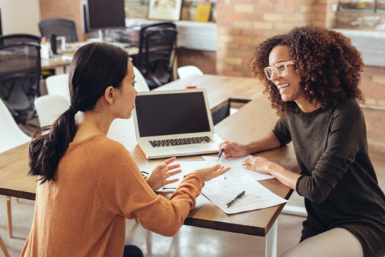 Photo of two women sitting in an office, at a table, talking