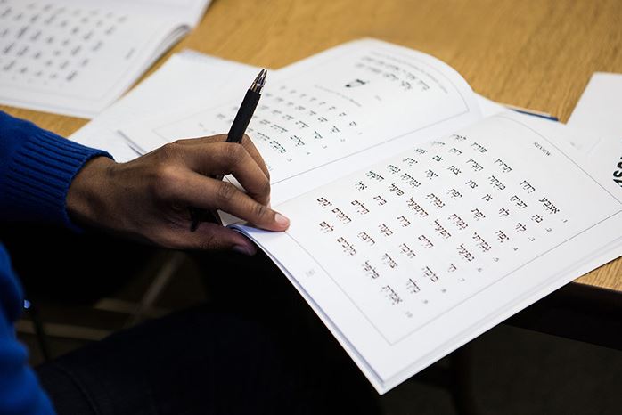 A hand holding a pen over a Hebrew workbook