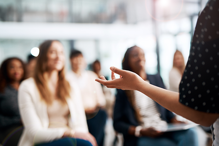 an image of a woman standing in front of a room of people, you see the arm of the woman and a few blurred out people in the background