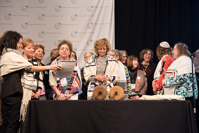 Rabbi Liz Hirsch standing with a group of women behind a table with a black table clothe; on top of table is a Torah and behind them is a white Women for Reform Judaism sign