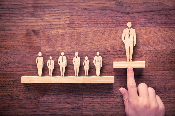 Photo of multiple wooden people figurines on two shelves; one figurine is on a shelf higher then the rest