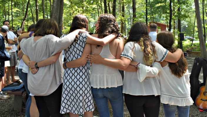 Teenage girls dressed in white with their arms around one anothers shoulders from behind 