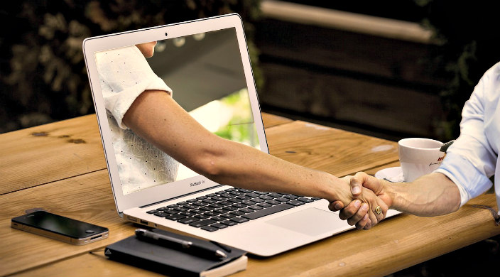 A mans arm reaches out through a computer screen to shake the hand of the man using the computer