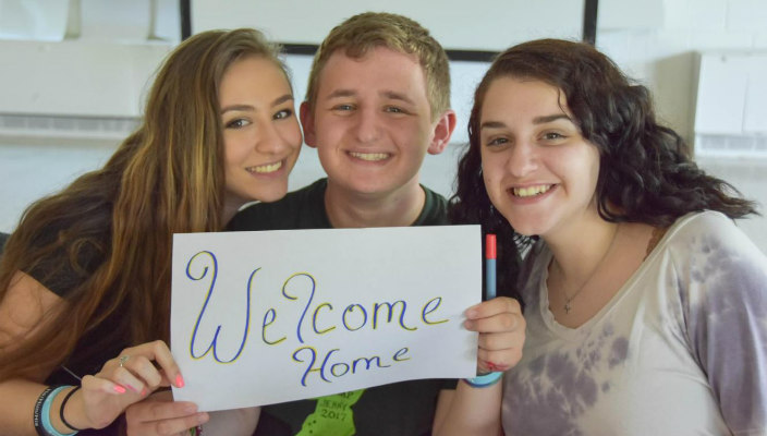 Three smiling teenagers holding a sign that reads WELCOME HOME