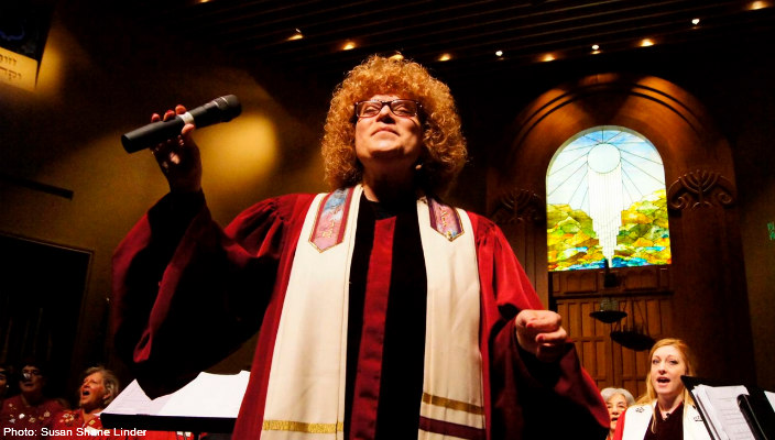 Closeup of a female rabbi with curly blond hair leading a congregation from the bimah with her arms open wide while wearing a prayer shawl