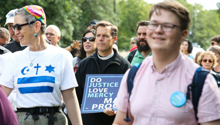 Three individuals as part of a larger group marching in some sort of event and carrying a sign that reads DO JUSTICE LOVE MERCY MARCH PROUDLY