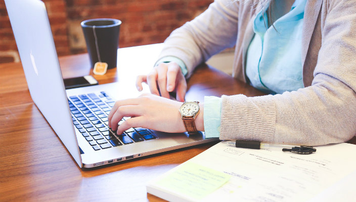 Closeup of a persons hand resting on a mouse as they sit at a laptop with a mug of coffee nearby as if working 