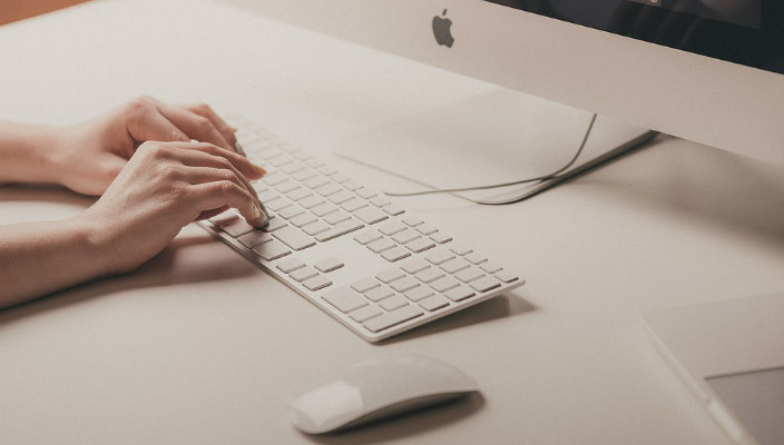 Caucasian womans hands on a white Apple keyboard with a mouse next to her and a monitor in front of her