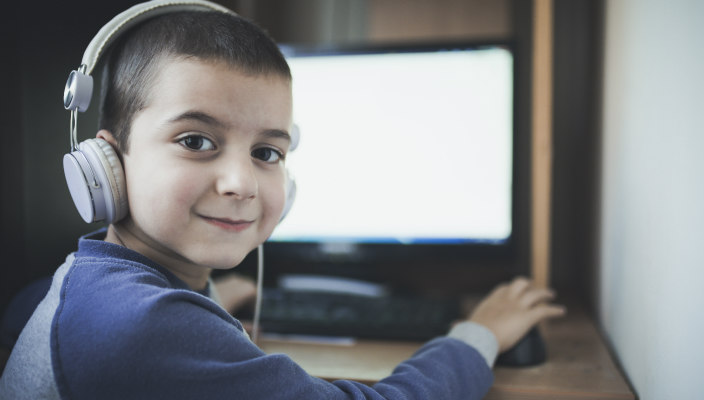 Boy wearing headphones and looking over his shoulder from a desk with a computer