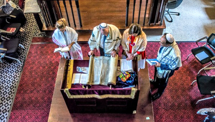Aerial view of four individuals in Jewish ritual garb standing at a bimah over an unrolled Torah scroll 