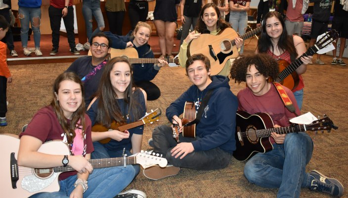A group of high school students sitting on the floor, each holding a guitar