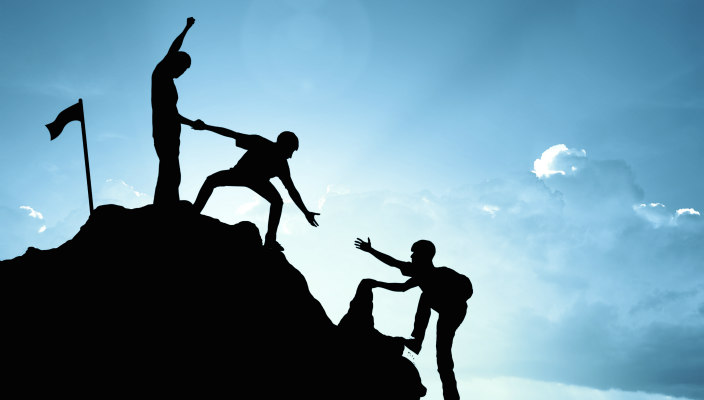 Three people in shadow helping each other ascend to the top of a mountain