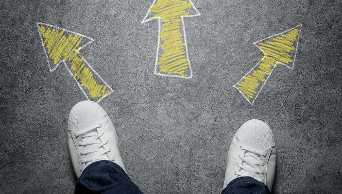 Pair of feet in white sneakers and blue pants seen from above, with three chalk-drawn arrows in front: one pointing left, one pointing right, and one pointing straight ahead