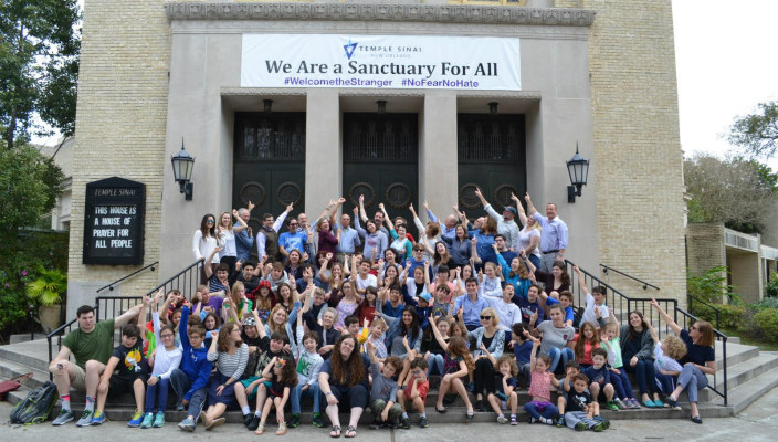 Large group of smiling people under a synagogue banner reading WE ARE A SANCTUARY FOR ALL