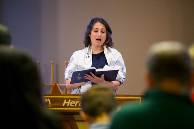 an iamge of a female cantor wearing a tallit, holding a prayer book and singing