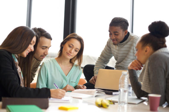 Photo of mulitple diverse people sitting around a conference table talking and looking at laptop