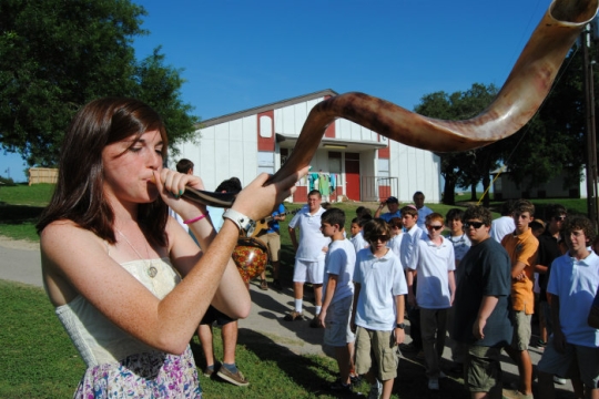 Teen girl blowing a shofar while a group looks on in front of a large outdoor structure as if at summer camp
