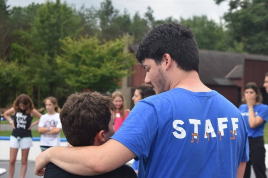 A mans back wearing a blue STAFF shirt with his arm around a young camper