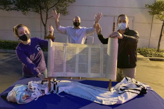 Three masked individuals holding up an unrolled Torah scroll on a table set outdoors