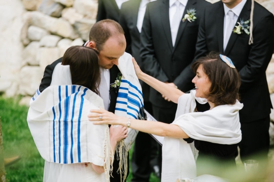 Bride and groom stand in front of a rabbi and are wrapped in a tallit