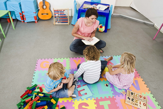 an image of a teacher sitting on a colorful mat with a book in her hands and three toddlers sitting in front of her
