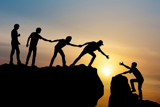 Photo of the silhouette of four people trying to help a fifth person cross a gap in between two rocks
