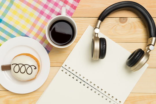 Photo of headphones, a cup of coffee, a pastry and a notebook on top of a wooden table