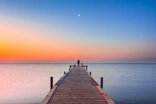 Photo of a person standing at the end of a dock on a body of water with the sun setting
