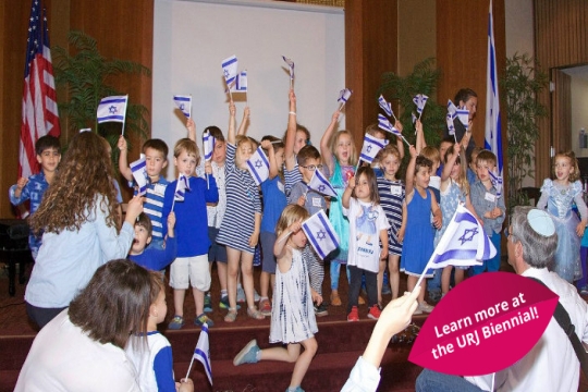 Young children waving Israeli flags on the stage in a synagogue social hall