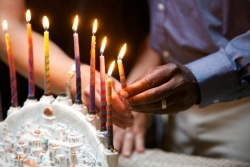 A womans white hand and a mans black hand hold a Hanukkah candle together as they light a Hanukkah menorah