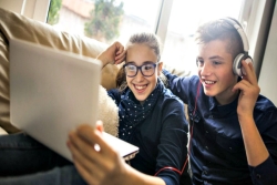 Two smiling teenage siblings sharing headphones and watching a laptop screen while sitting on a couch together