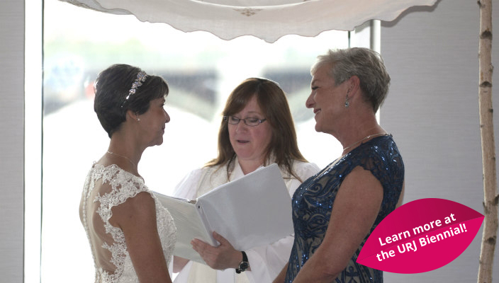 Two women holding hands under a chuppah as a rabbi officiates over their wedding ceremony