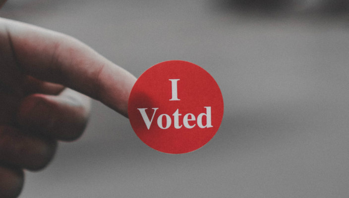 Finger holding a red and white sticker that reads I VOTED